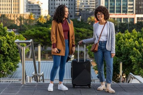 Two friends chatting and smiling with rolling suitcase on city terrace