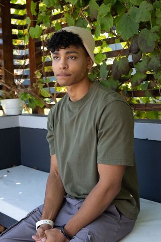 African american man sitting on patio bench wearing olive t-shirt and beige beanie