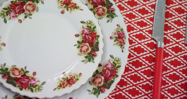 A floral-patterned plate is paired with a red-handled knife on a tablecloth with a red and white design, with copy space. The setting suggests a mealtime scenario, in a domestic or celebratory context.