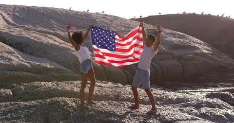 Young biracial couple celebrating with american flag on beach rocks