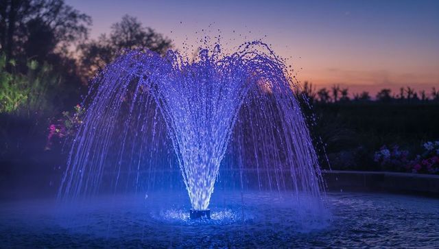 Glowing blue-purple fountain in dusk garden casting arcing jets and mirrorlike reflections
