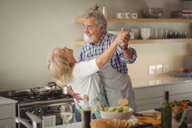 Senior couple dancing joyfully in their kitchen at home. They are smiling and enjoying each other's company, showcasing a happy and loving relationship. The kitchen is modern and well-lit, with various kitchen items and food on the counter. This image can be used for promoting healthy and active senior lifestyles, retirement living, family bonding, and romantic relationships in later life.