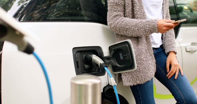 Woman leaning against electric car while charging it, using smartphone. Can be used for topics related to green energy, sustainable transportation, modern technology, environmental awareness, and eco-friendly lifestyle.
