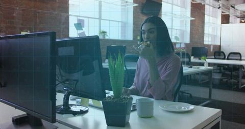 Professional Woman Eating Sandwich at Dual Computer Monitors in Modern Office