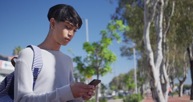 A young man is checking his smartphone outdoors on a sunny day. The man is holding the phone with both hands and is standing near trees and leafy surroundings under clear blue skies in casual attire including a gray shirt and backpack. Useful for concepts related to technology, communication, mobile usage, youth lifestyle, outdoor activities, and sunny weather.