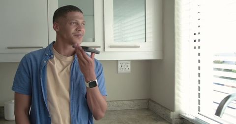 Young man using smartphone voice assistant in bright modern kitchen near window