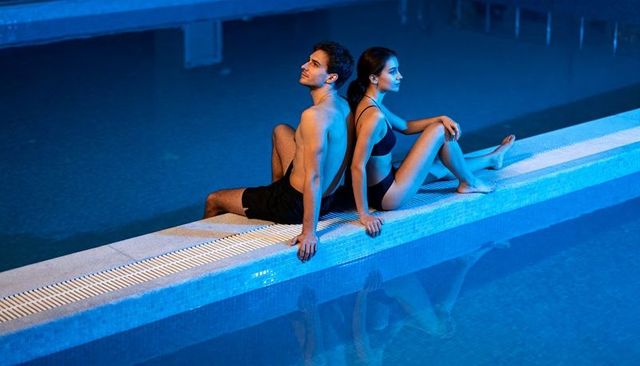 Couple sitting back-to-back on pool edge at night blue mood lighting reflective water
