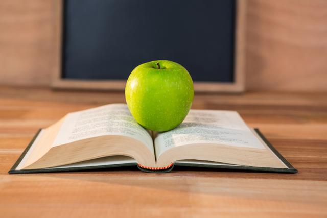 Green apple placed on an open book on a wooden table, with a blackboard in the background. Ideal for educational content, back-to-school promotions, healthy eating campaigns, and academic articles.