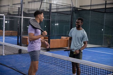 Two men chatting at net on indoor padel court holding rackets and ball casual sport
