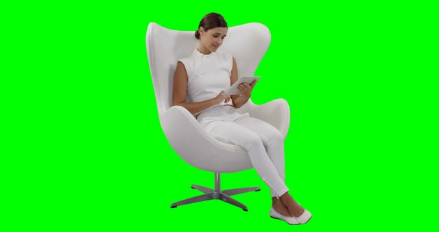 Businesswoman in white outfit using tablet while sitting in a modern chair with green screen background. Ideal for use in corporate presentations, technology-related content, remote work, and digital communication themes. Suitable for creating customizable backgrounds or integrating into various marketing materials.