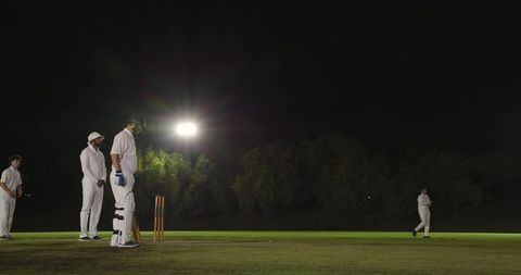 Night cricket match under floodlights with batsman at crease and club teammates waiting