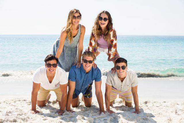 Young friends enjoy a sunny day at the beach by forming a human pyramid on the sand. They are dressed in casual summer clothing and sunglasses, conveying a sense of fun and camaraderie. This image is ideal for promoting summer vacations, beach activities, friendship, and outdoor fun.