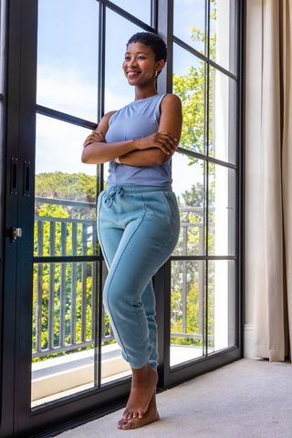 Sunlit relaxed woman standing barefoot by balcony door in casual loungewear