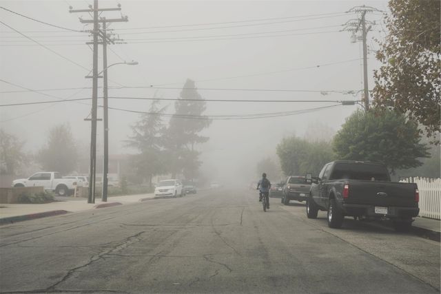 Depicts a misty morning on a suburban street with a single cyclist riding down the road and parked cars on the sides. The fog creates an eerie and calm atmosphere, obscuring the details in the distance. Power lines crisscross above, adding to the urban feel. Ideal for illustrating themes of solitude, calmness, suburban life, and weather-related conditions in urban areas.