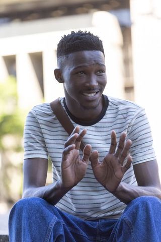Smiling young man gesturing while sitting on urban ledge wearing striped t-shirt and crossbody bag