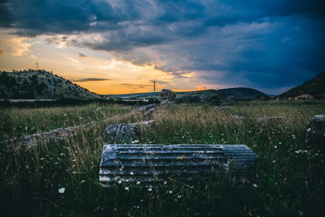 Broken columns and ruins lie scattered in a grassy field, under a sunset sky with dramatic clouds. Ideal for themes of history, archaeology, outdoor discovery, and travel brochures emphasizing historic tourism or sunset landscapes.