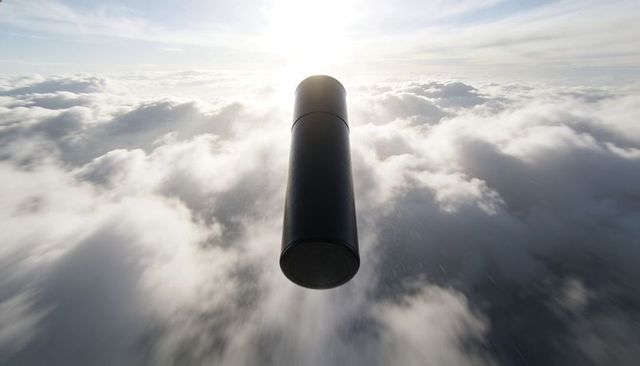 Dark metallic canister descending above sunlit cloud deck, backlit silhouette motion