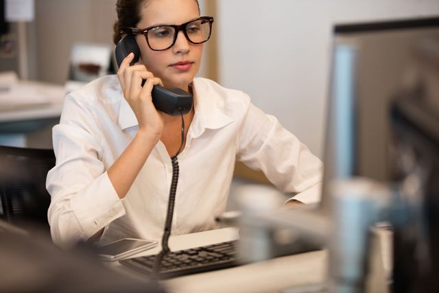 Businesswoman in white shirt using telephone while working at desk with computer in modern office. Ideal for illustrating corporate communication, professional work environments, business technology, and office productivity.