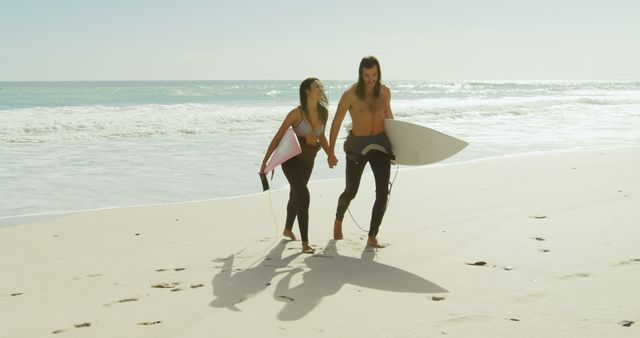 Caucasian couple enjoys a beach day, with copy space. They're walking along the shore, carrying surfboards and ready for a surfing session.