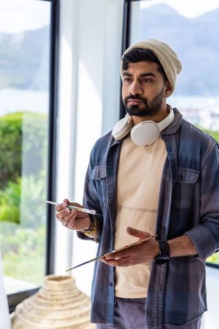 Asian man painting by large window wearing beanie and headphones in bright lakeside studio