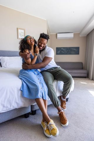 Laughing african american couple hugging on bed sharing phone in modern cozy bedroom