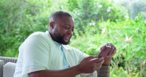 African american man relaxing outdoors using smartphone on wicker chair in garden