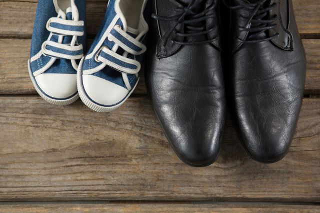 Image shows a pair of adult black formal shoes next to a pair of child's blue sneakers on a wooden surface. This visual contrast highlights the bond between parent and child, making it ideal for themes related to family, parenting, growth, and lifestyle. Perfect for use in advertisements, blogs, and articles focusing on family relationships, parenting tips, and lifestyle choices.