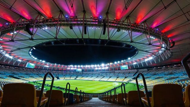 Empty floodlit stadium interior with circular roof ring, yellow seats and lush soccer pitch