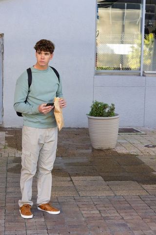 Teen standing on city sidewalk checking phone while holding paper lunch bag and backpack