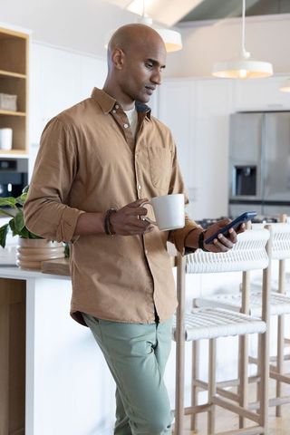 African american homeowner leaning on island holding mug and checking phone