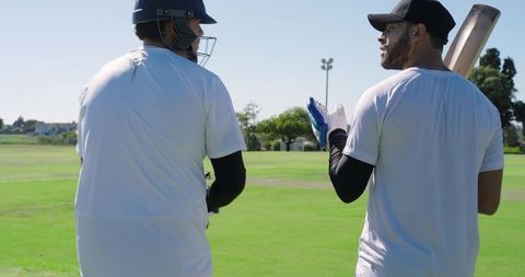 Walking male cricket teammates carrying bats and spare helmet toward wickets on grass
