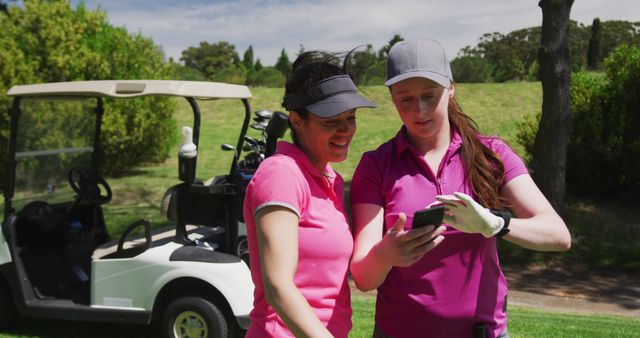 Female golfers are checking their score on a smartphone during a match. They are standing near a golf cart that is loaded with golf clubs, adding to the sporty context. This image can be used for websites or advertisements focused on women's sports, golf courses, outdoor activities, and teamwork. It is also suitable for illustrating articles about golf, apps for tracking sports scores, and women’s participation in sports.