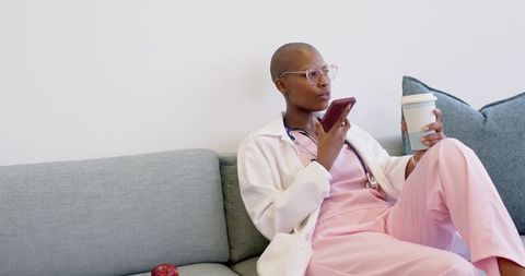 Nurse taking break holding coffee and smartphone on sofa in pink scrubs with stethoscope