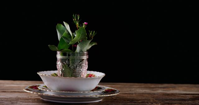 A vintage glass with green leaves and a pink flower is placed in an ornate saucer on a rustic wooden surface, with copy space. The dark background accentuates the elegance of the table setting and the simplicity of the plant arrangement.