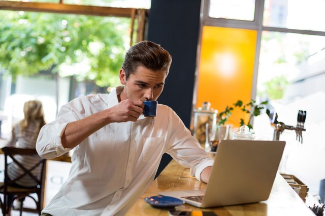 Young man drinking coffee while working on laptop in a modern cafe. Ideal for illustrating remote work, freelancing, urban lifestyle, and casual business settings.