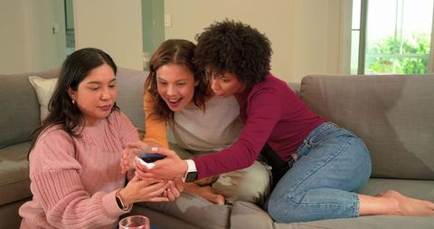Friends sharing smartphone and laughing on cozy sofa at home, candid lifestyle moment