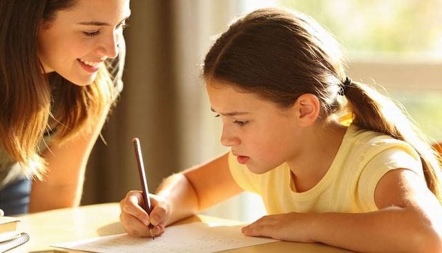 Young girl concentrating on homework with smiling tutor at sunlit desk