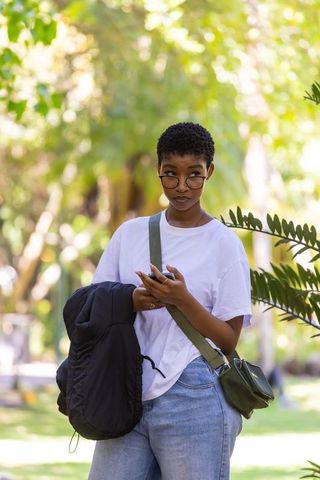 Young woman wearing glasses checking smartphone while holding jacket in sunlit park