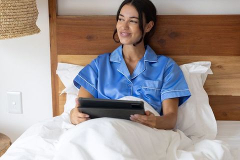 Woman relaxing in blue satin pajamas on bed holding tablet cozy minimalist morning