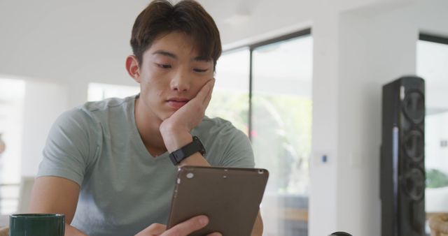 Bored asian male teenager using tablet and sitting in living room. spending time alone at home.