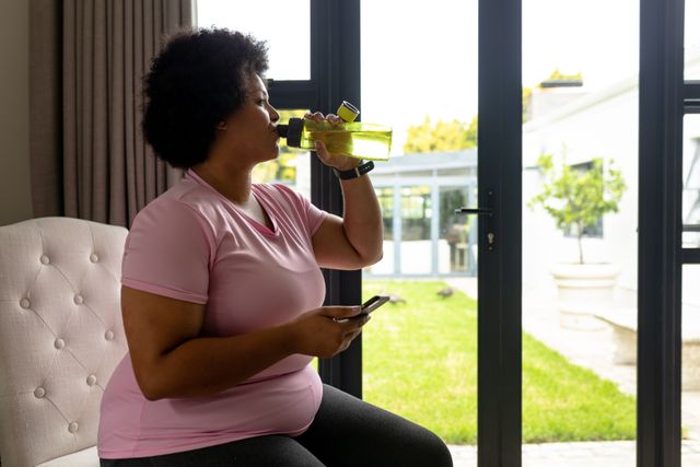 African american mid adult woman drinking water while sitting with smart phone on chair at home. unaltered, wireless technology, drink, fitness and active lifestyle concept.