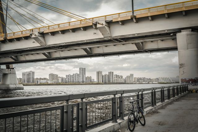 Bicycle resting against fence on waterfront under large concrete overpass with urban cityscape in background. High-rise buildings and river visible under cloudy sky. Ideal for illustrating urban commutes, cityscapes, contemporary life, transportation.
