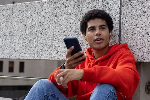 Teen sitting on rooftop wearing red hoodie holding smartphone and earbuds, casual style