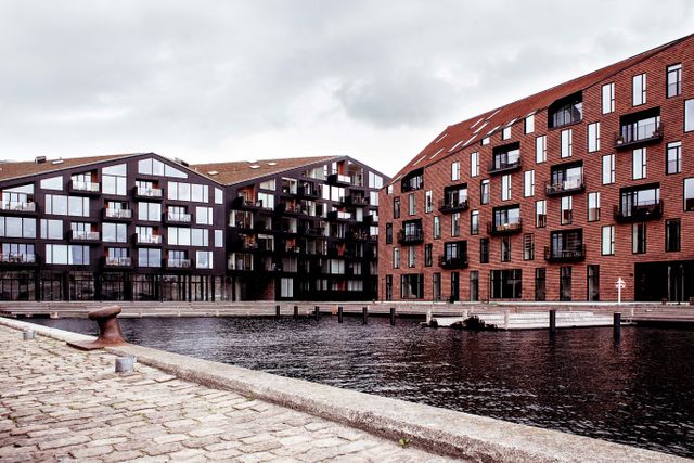 The image showcases modern Scandinavian apartment buildings along a waterfront on a cloudy day. The architecture features large windows and multiple balconies, with building facades in a mix of dark and reddish tones. This image is ideal for use in topics related to urban living, real estate, architecture, and contemporary residential development.