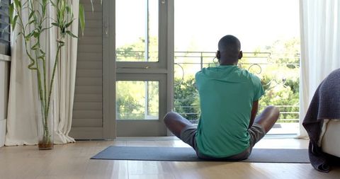 Man in 30s sitting on yoga mat facing balcony, wearing earbuds, enjoying sunlight in apartment
