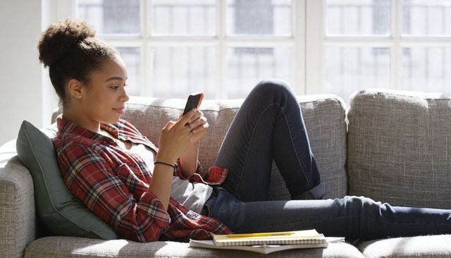 Young woman relaxing on sunlit sofa texting on smartphone with notebook nearby