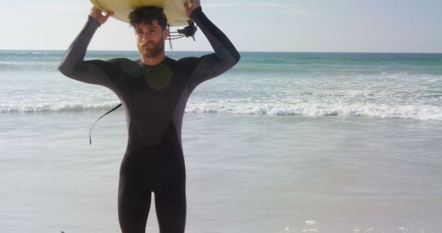 A young Caucasian man in a wetsuit carries a surfboard on a sunny beach, with copy space. His relaxed posture and the serene ocean backdrop suggest a leisurely day of surfing ahead.