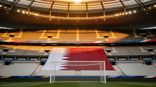 Giant qatar flag draping central stands in modern stadium at golden hour with empty seats
