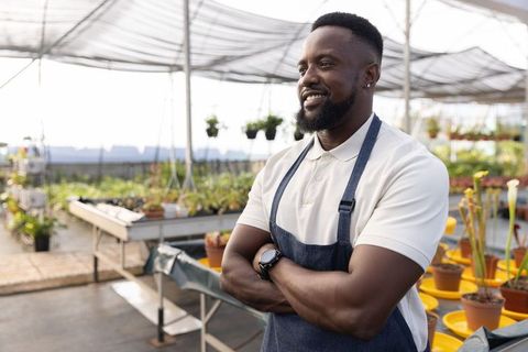 Smiling gardener standing in greenhouse with crossed arms wearing apron and polo