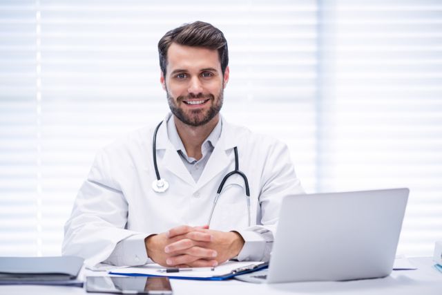 Male doctor sitting at desk in clinic, smiling confidently. Stethoscope around neck, laptop and medical documents on desk. Ideal for healthcare, medical services, professional consultation, and patient care promotions.